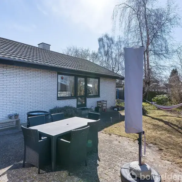 En terrasse foran et hvidt murstenshus. Sorte havemøbler står omkring et bord. En parasol er pakket ind. Bagved ses en have med en hængekøje og nogle træer.