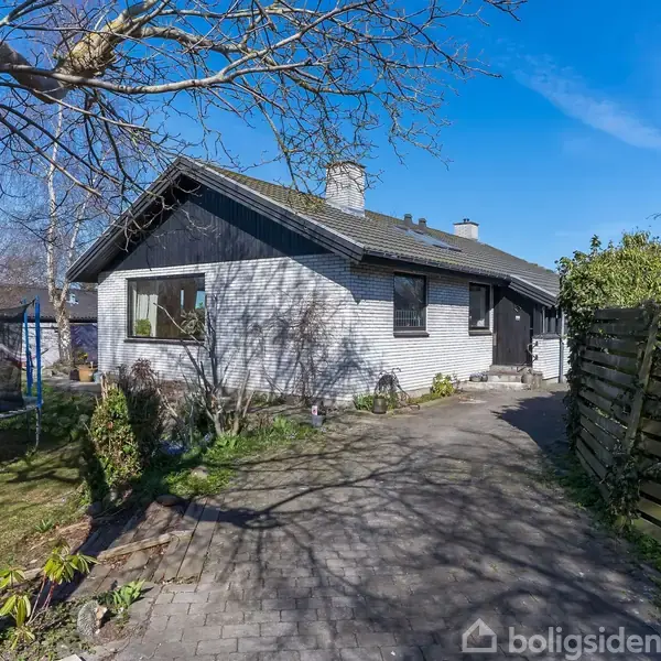 Et gråhvidt hus med mørkt tag står på en flisebelagt indkørsel omgivet af have og træer. En trampolin ses til venstre, og et hegn begrænser arealet til højre.