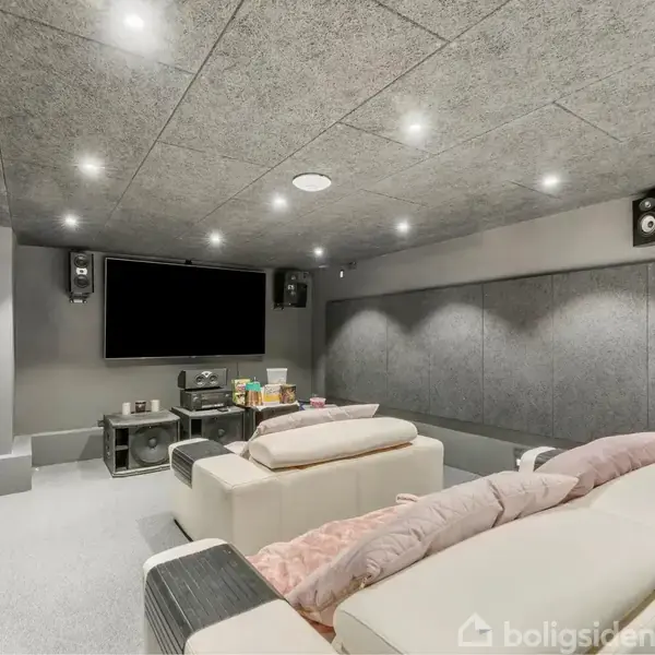 Blød sofa vender mod en stor TV-skærm i et moderne hjemmeteater. Bløde tæpper og surroundhøjttalere omkranser rummet, der er dækket af lydabsorberende paneler og dæmpet belysning.