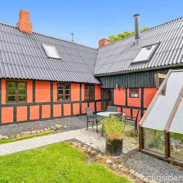 Rødt bindingsværkshus med sorte bjælker, har terrakottatag og flere vinduer. Der er et udendørs siddeområde med bord og stole i en lille have med grøn græsplæne og flisestier.