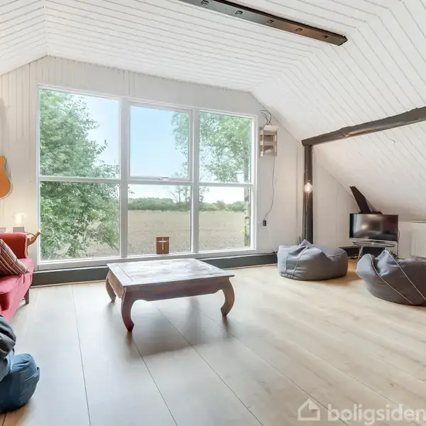 Stue med stort vindue viser udsigt til naturen, rød sofa med pude, guitar på væggen, lavt bord, tre sækkestole og lille TV i et lyst rum med skråt loft.