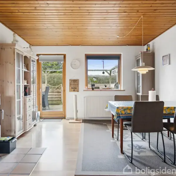 En brændeovn med flammer står i en lys stue med trægulv. Et spisebord er dækket med en mønstret dug, og der er udsigt til en terrasse gennem glasdøre.