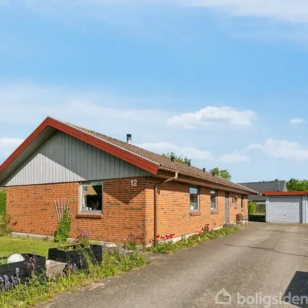 En rød murstensvilla med skifertag står i en indkørsel, omkranset af en grøn have med blomsterbede. To garager ses i baggrunden. Nummer 12 er synligt på husets facade.