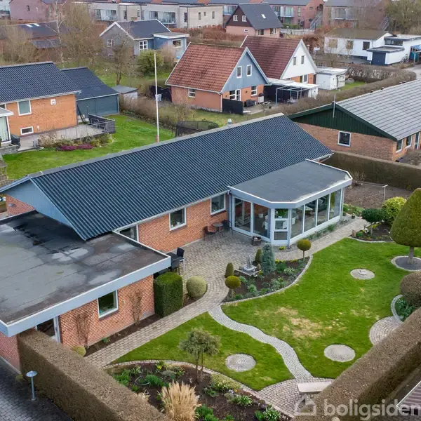 Et rødt murstenshus med sort tag hviler i en velholdt have med stier og buske. Huset har en tilstødende udestue, omgivet af naboer i et boligområde.