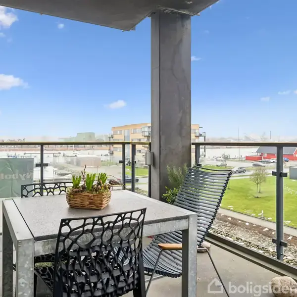 Altan med et bord og stole står stille. I baggrunden ses en grøn græsplæne og moderne bygninger under en blå himmel med få skyer.