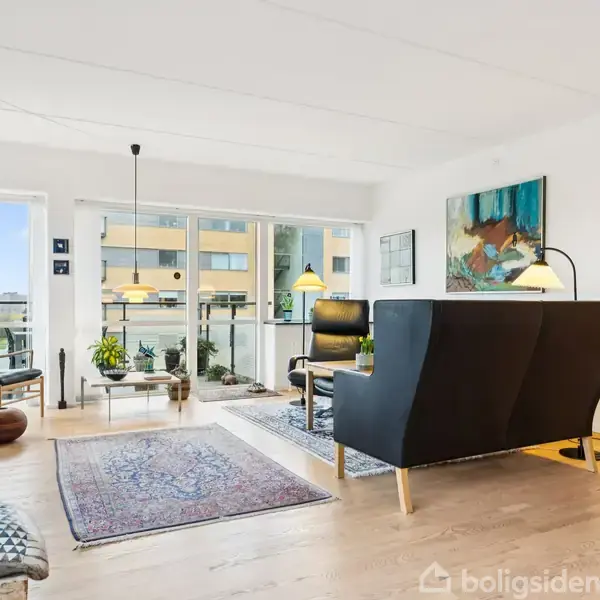 Stue med sort sofa og træmøbler, dekoreret med planter og kunstværker, mod glasskydedøre til balkon. Lyst trægulv og tæpper skaber en moderne og indbydende atmosfære.