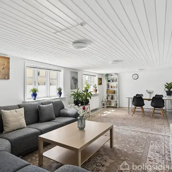 Grå hjørnesofa med puder står i stue, foran et lyst sofabord med blomster. Spisebord og stole i baggrunden, planter og kunst på hvide vægge, tæppe på gulvet.
