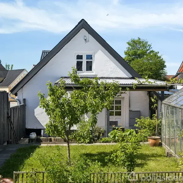 Hus med hvid facade, lille vindue og sort tag gør. I en grøn have med et drivhus til højre og et hegn til venstre om sommeren.