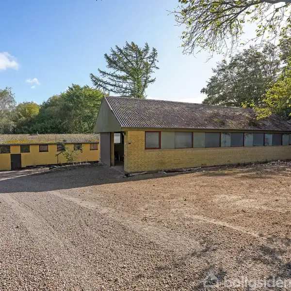 En gul bygning med vinduer står på en grusvej omgivet af træer og buske. Yderligere en mindre bygning ses til venstre, delvist synlig gennem indgangen.