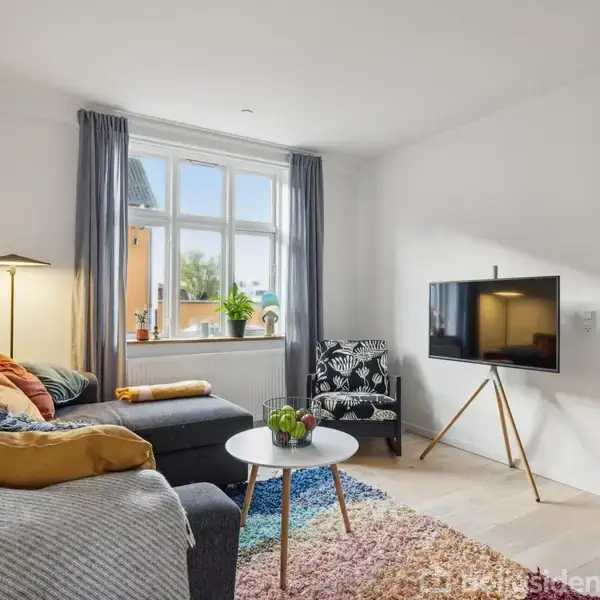 En grå sofa med farverige puder står i en stue med et tæppe. Et fladskærms-TV er monteret på en stander. Der er et vindue med blå gardiner og en stol.