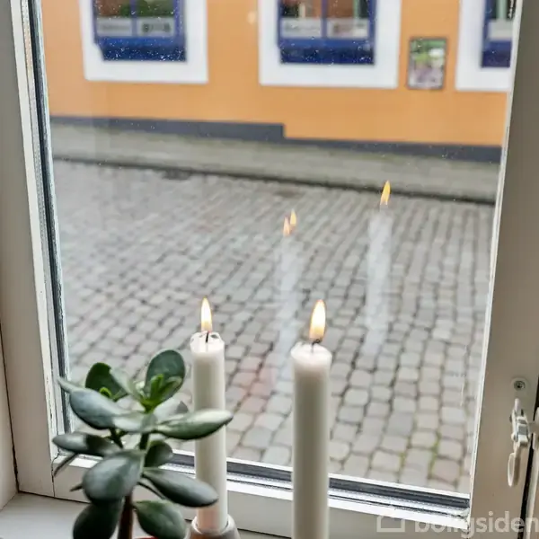 Tre stearinlys brænder i vindueskarmen med en plante ved siden af. Udenfor ses brosten og en gul bygning med blå rammer.