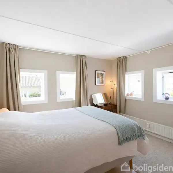 Soveværelse med en dobbeltseng, som har et hvidt sengetæppe og en blå plaid. Fire små vinduer med gardiner slipper lys ind. Et skrivebord med spejl og lampe står ved væggen.