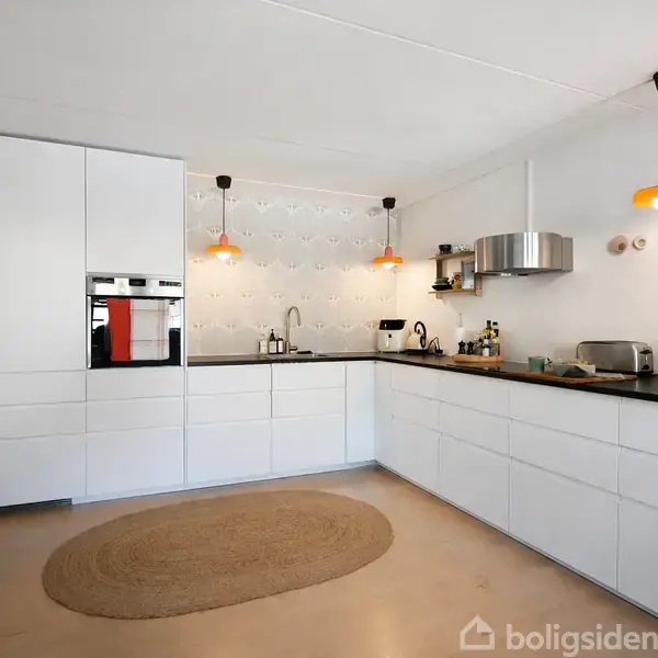 Køkken med hvide skabe og sort bordplade; to orange pendellamper hænger over bordpladen. Der er indbygget ovn, stålventilator og køkkenudstyr. Væggene har minimalistisk dekoration.