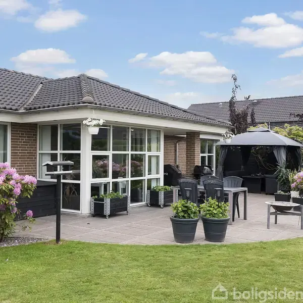 En villa med store vinduespartier og en terrasse med havemøbler. Omkring huset er der blomstrende buske og en velplejet græsplæne. En pavillon er opstillet på terrassen.