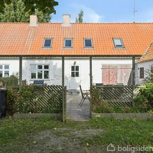 Rækkehus med rødt tag og hvide vægge står stille omkranset af en lille have med grønne buske. Der er en terrasse med et bord og stole foran huset. To affaldscontainere står i venstre side.