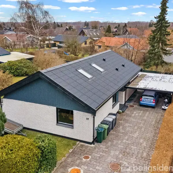 En moderne, hvid murstensvilla med mørkt tag står i en stille forstad. Bilen holder i carporten ved siden af. Omkring huset ses haver og træer. Nummer "97" på væggen.