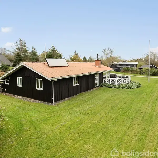 Sort hus med rødt tag står i en velplejet have med græsplæne. På taget er der en solfanger. Området er omgivet af træer og fælleshuse.