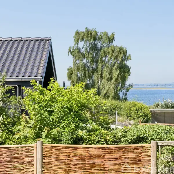 Et hus med sort tag ligger ved siden af tæt grøn beplantning og en flethegn. En stor træ top er synlig, og der er udsigt til havet i baggrunden.