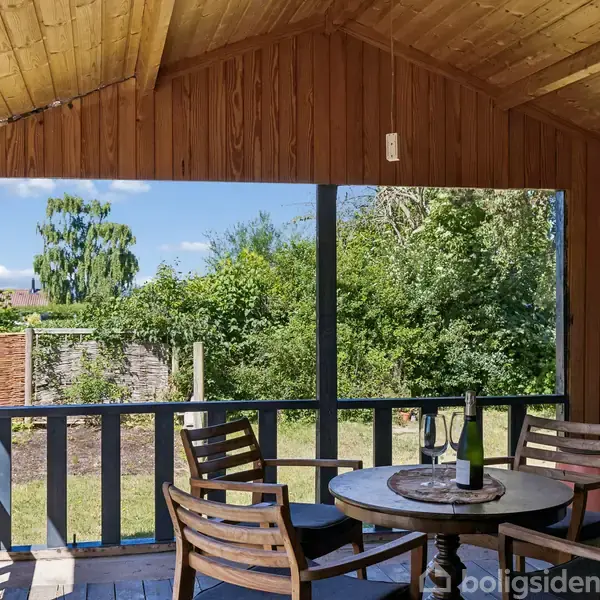 Træmøbler står på en overdækket terrasse med udsigt til en frodig have. En flaske og et glas er på bordet. Sollys filtreres gennem træloftet.