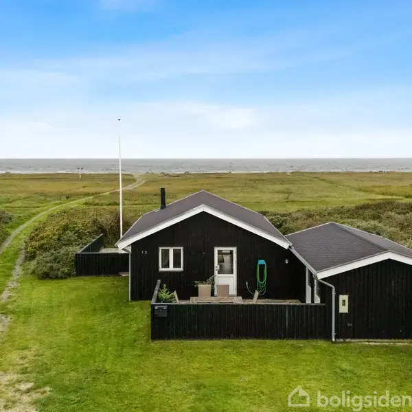 Et sort, træsommerhus står stille på en grøn græsplæne med udsigt til havet i baggrunden. En grussti fører op til huset, omgivet af lav vegetation.