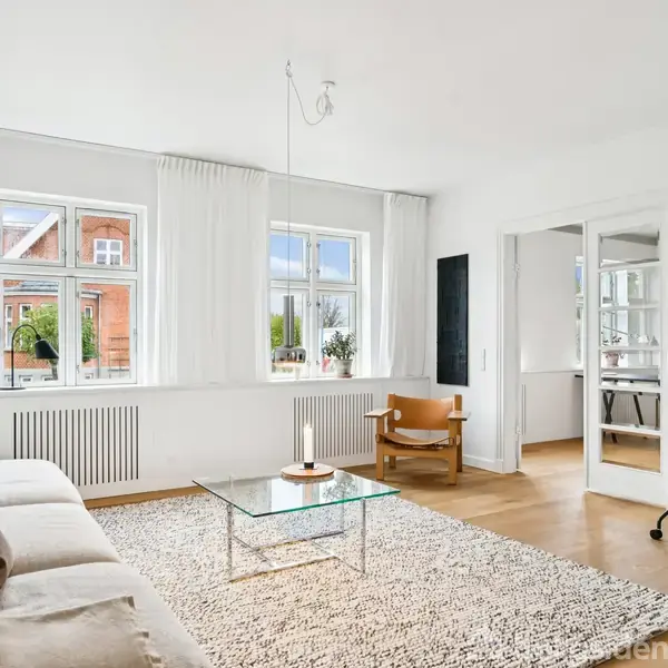 En lys stue med en beige sofa og en glassofabord på et tæppe. Store vinduer slipper naturligt lys ind. Et hjørne indeholder en lænestol og fjernsyn.