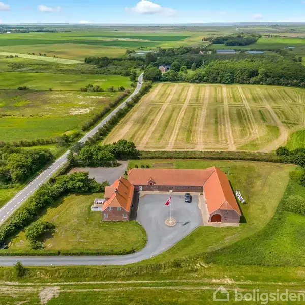 En gård med rødt tag og flagstang står i en stor, grøn mark. En lang vej løber forbi omgivet af marker og træer i det fjerne.