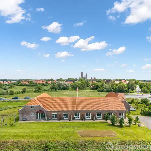 Bygning med rødt tegltag står stille på en grøn mark, omgivet af veje og marker. I baggrunden ses en by med kirketårne under en blå himmel med skyer.