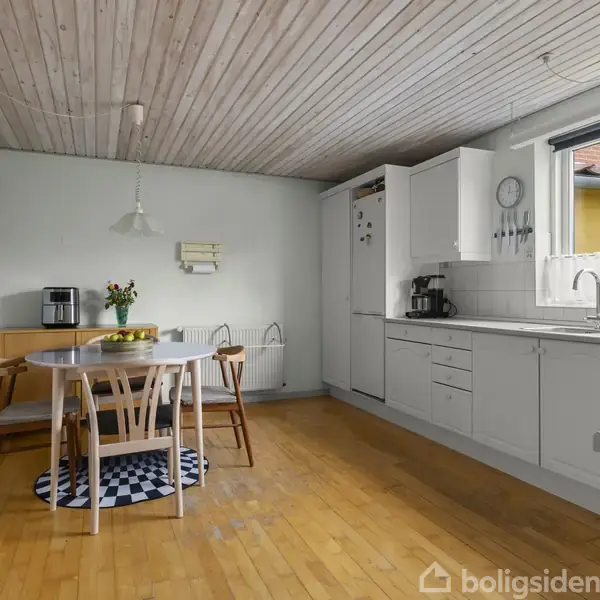 Køkkenbord med fire stole står på et trægulv. Bordet er dækket med blomster. Indrammet af hvide køkkenskabe og et stort vindue med udsigt til et gult hus.