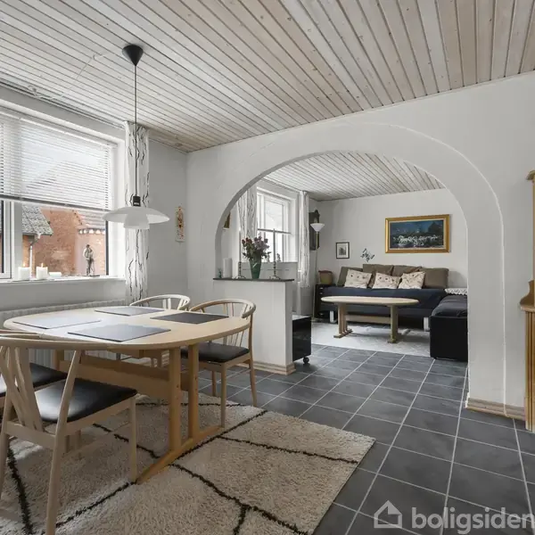 Et spisebord med stole står i en lys spisestue med store vinduer. Rummet har flisegulve og trælofter. I baggrunden ses en stue med sofa og billeder på væggen.