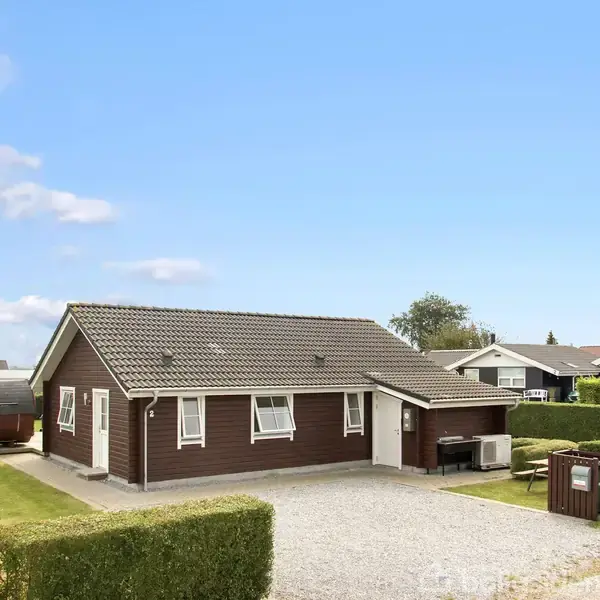 Et brunmalet hus står stille omgivet af en velplejet grusindkørsel og hække. Det er en solrig dag med blå himmel i et roligt beboelsesområde med flere lignende huse.