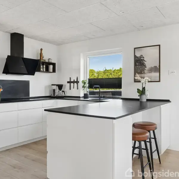 Et moderne køkken med hvide skabe og sort bordplade. En ø med to barstole foran. Et vindue i baggrunden viser grønne træer udenfor. En plakat hænger på væggen.