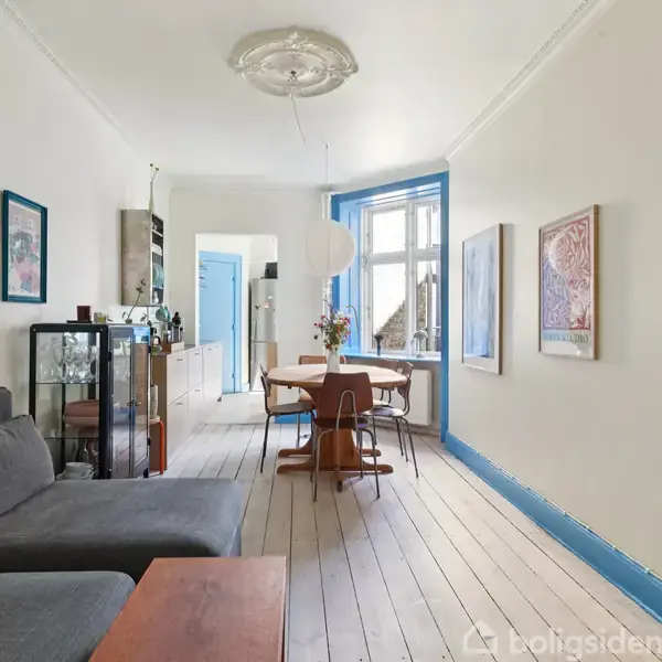 En stue med grå sofa, akvarieskab og spisebord. Store vinduer til venstre lader lys ind, mens vægkunst og en lysekrone pynter rummet. Blåt dørkarme og trægulv.