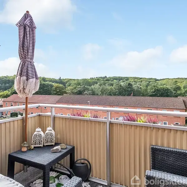 En altan med kurvemøbler og lanterner står stille, mens en udsigt til grønne bakker og et taglandskab strækker sig i baggrunden under en blå himmel.