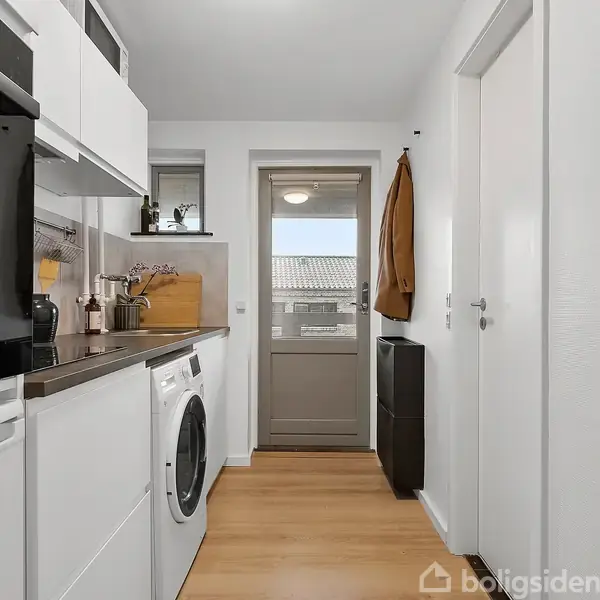 Vaskemaskine placeret i et moderne bryggers med hvide skabe, sorte bordplader og trælaminatgulv. En krukke, skærebræt og kaffemaskine står på bordet. En dør fører til ydersiden.