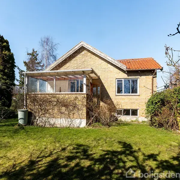 Et hus i gule mursten står stille i en grøn have med buske og træer omkring. På huset ses en overdækket terrasse med glasvægge. Klimaet virker roligt og solrigt.