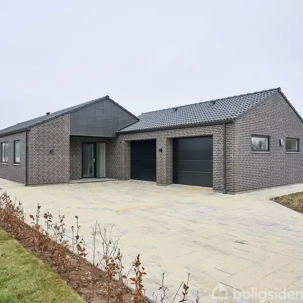 Et moderne, en-etagers, gråt murstenshus med fladt tag står på en åben grund. Foran huset er der en bred indkørsel med fliser, omkranset af en lav hæk.