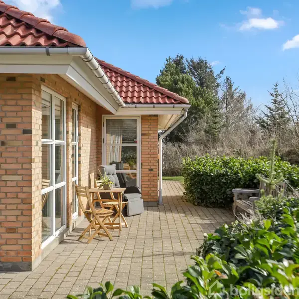 Murstenshus med rødt tag og store vinduer. Terrasse med havemøbler foran huset. Omgivet af buske med træer i baggrunden under en blå himmel.