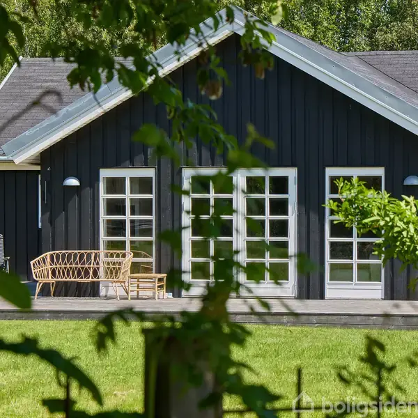 Et sort træhus med store glasdøre står i en grøn have. Foran huset er der en terrasse med en flettet sofa. Der er træer i baggrunden og forgrunden.