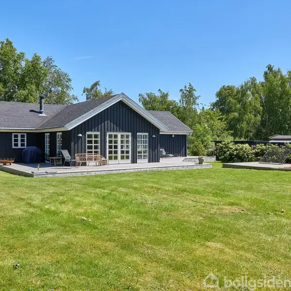 Et mørkt træhus står på en velplejet grøn græsplæne. Terrassen foran huset har udendørsmøbler og en grill. Omkranset af frodige træer og buske, giver det en rolig atmosfære.