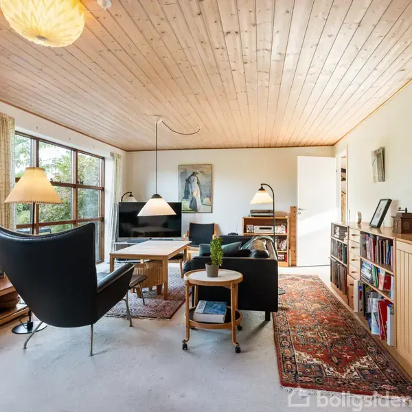 Stue med sorte lænestole og sofa arrangeret omkring et sofabord under hængelamper, omgivet af reoler og trævægpaneler. Store vinduer viser en grøn udsigt. Farverig tæppebelægning på gulvet.