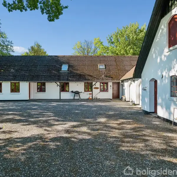 Hvide bygninger med røde døre og vinduer står omkring en gårdsplads med grus. Sollys skaber skygger fra træerne, der omkranser området.