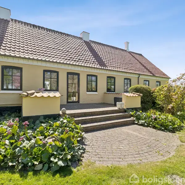 Hus med teglstenstag står stationært med trin fører til hoveddøren. Baggrunden inkluderer en græsplæne og et blomstrende træ under en klar himmel.