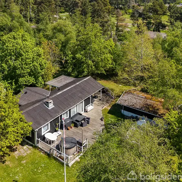 Et enkelt etplanshus med en mørk tag, omgivet af grønne træer i en skovklædt have, har en terrasse med havemøbler og et mindre skur ved siden af.