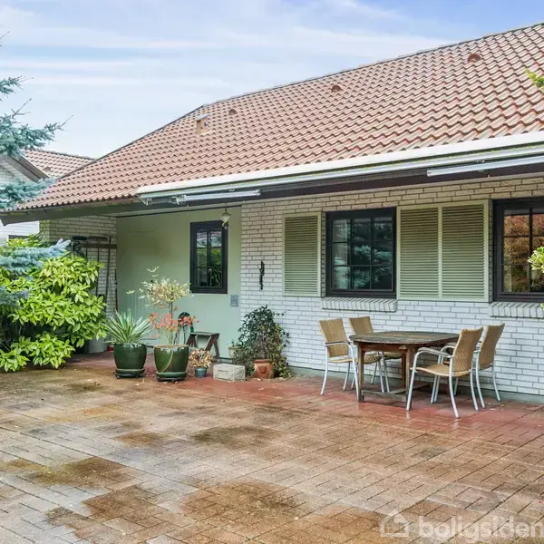 En flisebelagt terrasse med et rundt bord og fire stole placeret op ad en hvid murstensbygning. Omgivet af grønne buske og træer under en blå himmel.