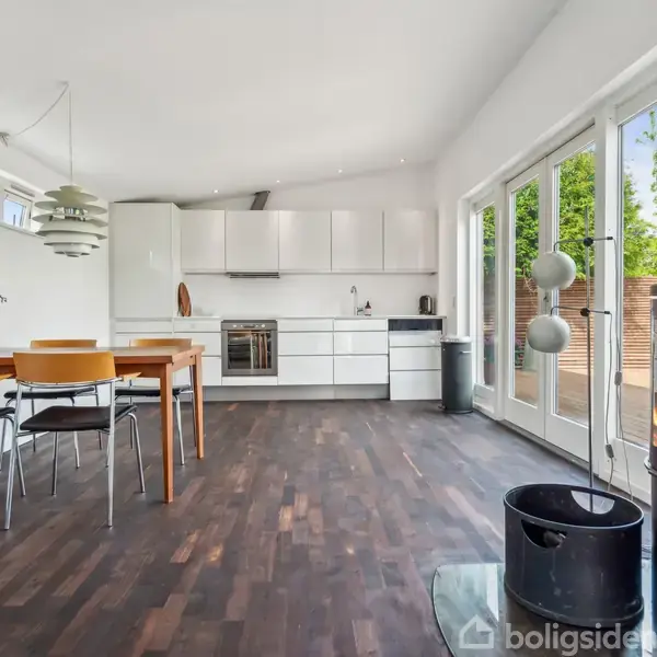 Et moderne køkken-alrum med hvidt køkkenskabe, bordplade og integreret ovn. Rundt spisebord med fire stole står ved siden af en pejs med ild. Store vinduer med udsigt til grønne omgivelser.