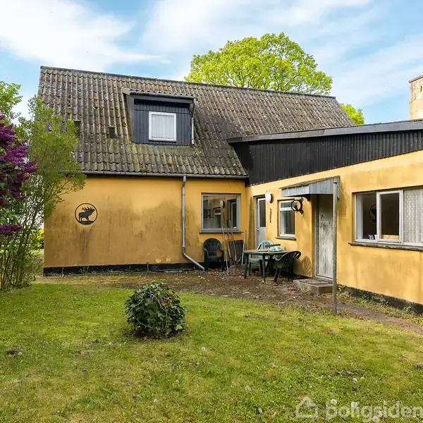 Et gult hus med sort tag har et lille terrassedæk med et bord og to stole foran. Græsplænen omgiver huset, og lilablomstrende busk til venstre.