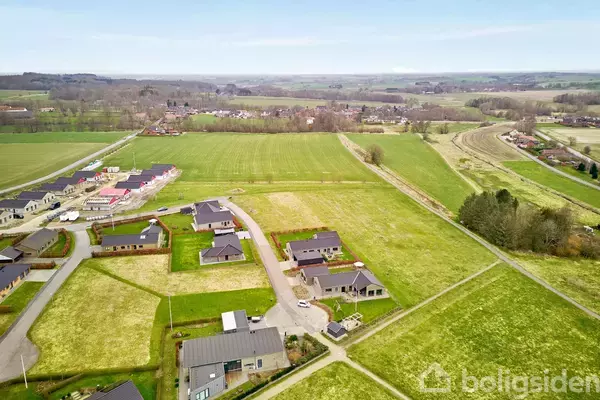 Landsbyhuse ligger spredt i et åbent, grønt landskab med marker og vejnet. I baggrunden ses en lille bebyggelse omgivet af skov og bakker.
