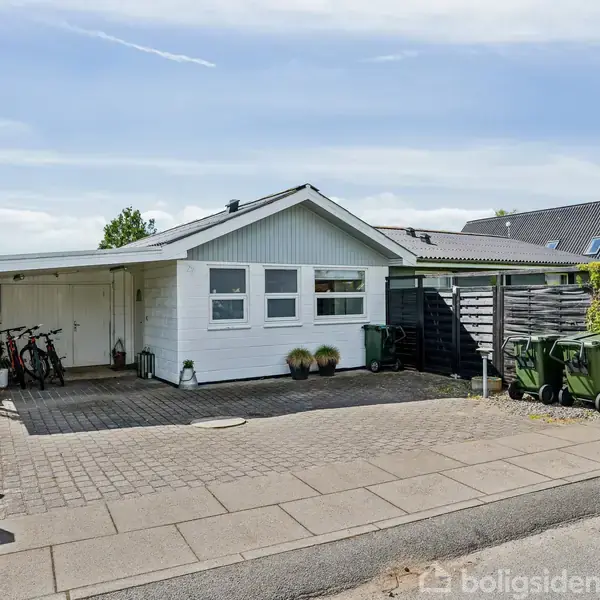 Hvidt hus står stille, foran parkeret cykler og skraldespande; det ligger i et villakvarter. Adressen "24" ses på postkassen.