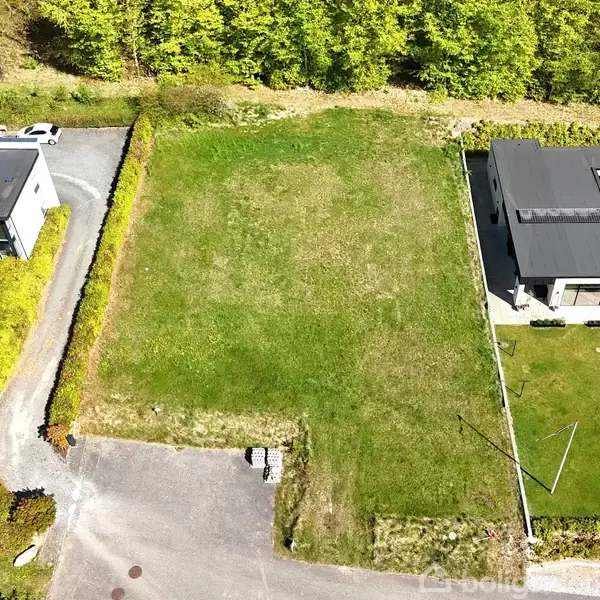 Byggegrund uden aktivitet omgivet af to moderne huse med grå tage, beliggende ved en asfalteret vej i et grønt område med træer i baggrunden.