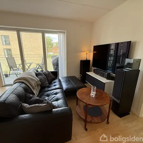 Sort lædersofa står foran en stor fladskærm-TV i en lys stue. Et rundt træbord holder et glas. Store vinduer leder ud til en balkon med stole.
